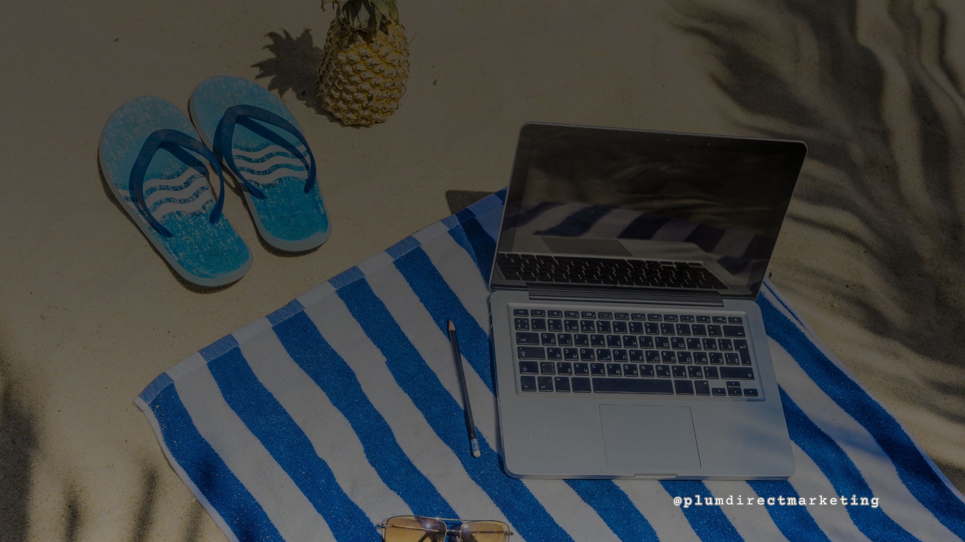 Laptop on striped towel at the beach with flip-flops and pineapple nearby, representing relaxed summer content planning for July Instagram marketing campaigns.